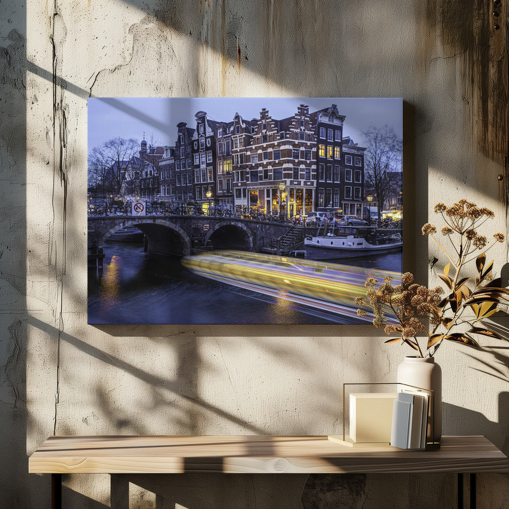 A long-exposure photograph of an Amsterdam canal at dusk, featuring a bright yellow light trail from a passing boat. In the background, illuminated historic canal houses line the water, and an arched stone bridge covered in bicycles crosses the canal. The entire image is presented in a silver frame. Print