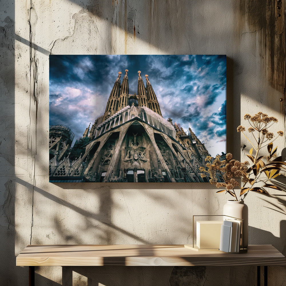 A dramatic, low-angle photograph of the Sagrada Familia in Barcelona, with its ornate stone facade and towering spires reaching up into a turbulent, dark blue and cloudy sky. The perspective emphasizes the grand scale and intricate detail of Gaudí's architecture. Print