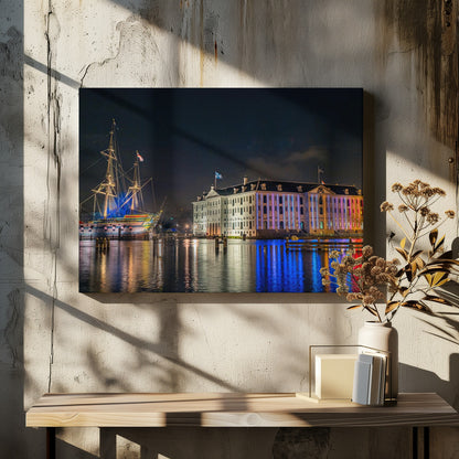 A vibrant nighttime photograph of the National Maritime Museum and a replica of the East Indiaman Amsterdam ship in a harbor. Both are illuminated with colorful lights that cast beautiful reflections on the dark water. Wall Art