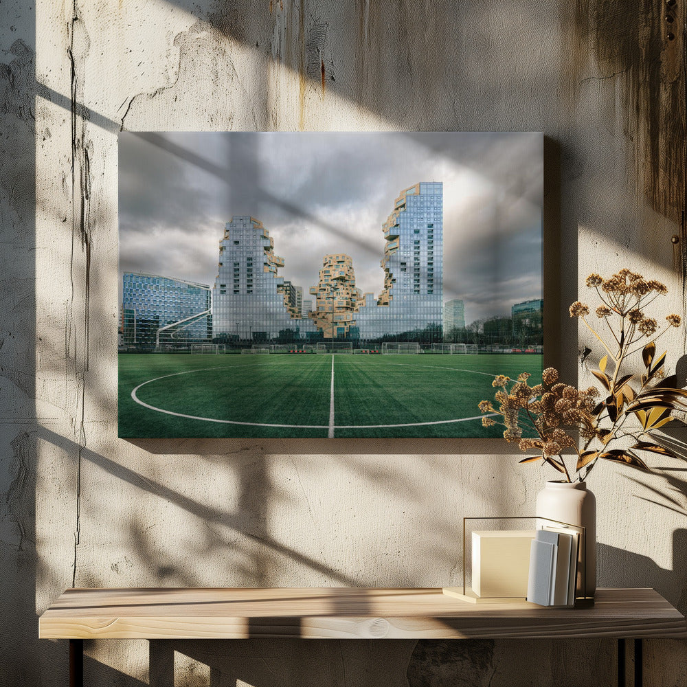A wide-angle view from the center of a lush green soccer field looking towards a striking modern architectural complex with uniquely shaped glass and tan buildings under a dramatic, cloudy sky. Decor