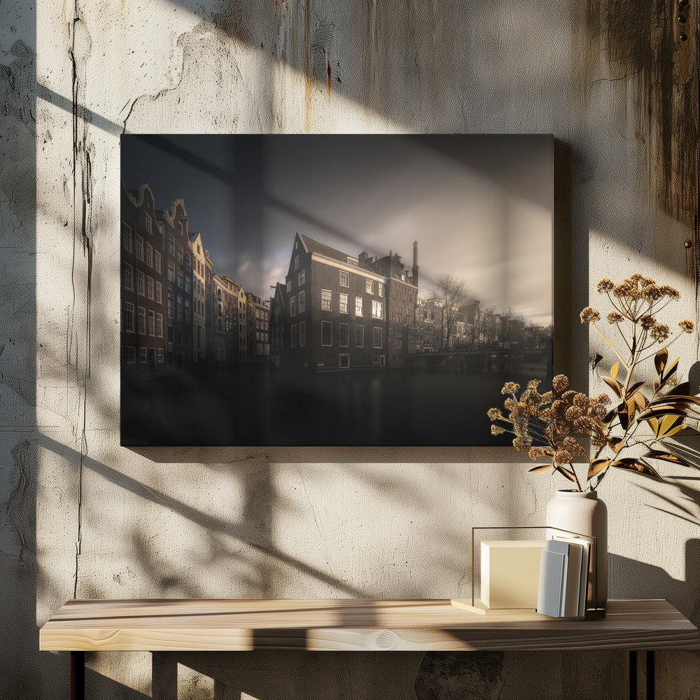 A moody, atmospheric photograph of historic canal houses in Amsterdam, viewed from the water. The scene is dark, with a dramatic, cloudy sky and a soft glow on the horizon, while some windows in the dark brick buildings are lit from within. The image is bordered by a silver frame. Wall Art