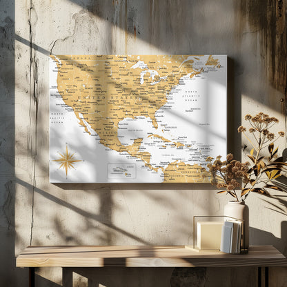 A stylized map of North and Central America and the Caribbean, featuring gold-colored landmasses on a light gray, textured background. Countries, states, and major cities are labeled in black text. A compass rose is shown in the lower-left corner. The entire map is enclosed in a light gray frame. Decor