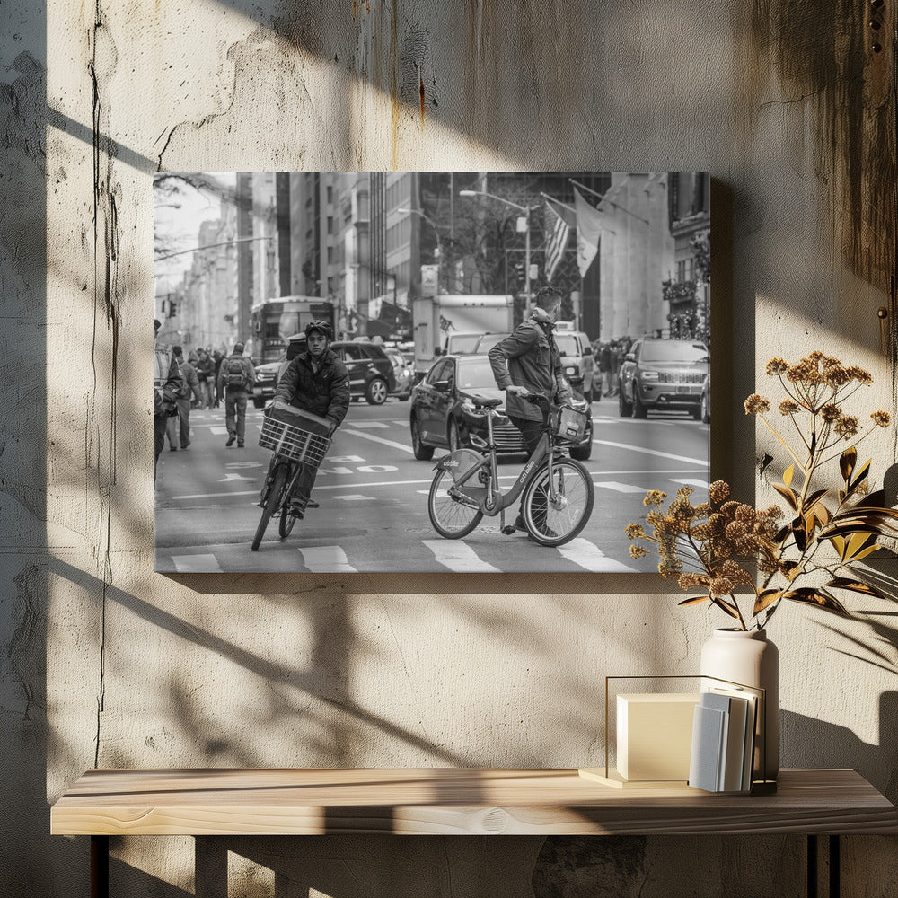 A black and white street photograph of a busy city intersection, framed in silver. In the foreground, two men are crossing with their bicycles amidst traffic, with one cyclist looking directly at the camera. Tall buildings and other pedestrians line the background, capturing a moment of everyday city life. Artwork