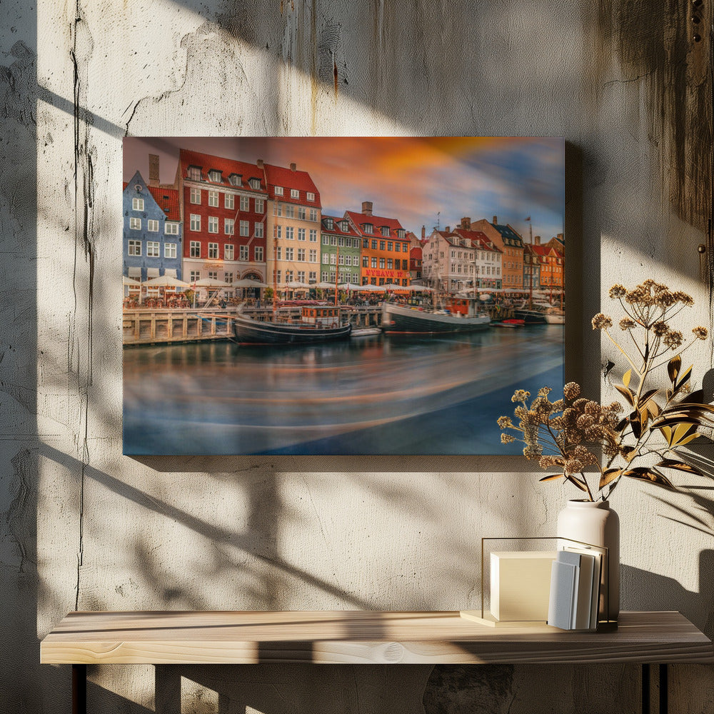 A long-exposure photograph of the colorful Nyhavn harbor in Copenhagen at sunset. Historic, brightly colored buildings line the canal, where two traditional wooden boats are moored. The sky is a dramatic blend of orange and blue, and the water is blurred, showing streaks of reflected light. Decor
