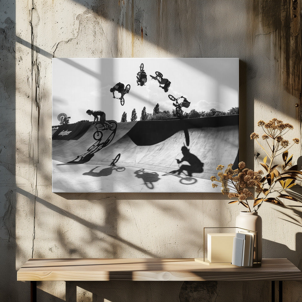 A dynamic black and white action shot showing the progression of a BMX rider doing a backflip off a large ramp. Multiple exposures capture the rider at different points in the air, creating an arc of motion against a bright sky. Strong shadows are cast on the ramp below. Print