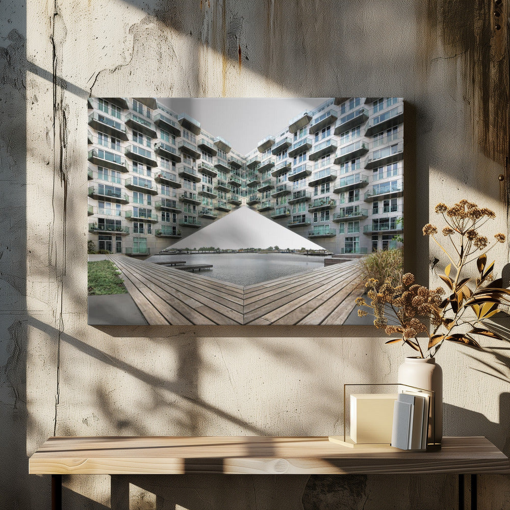 A symmetrical, mirrored photograph of a modern V-shaped apartment complex with numerous glass balconies, overlooking a wooden deck and a calm body of water under a grey sky. The image is presented within a silver picture frame. Artwork