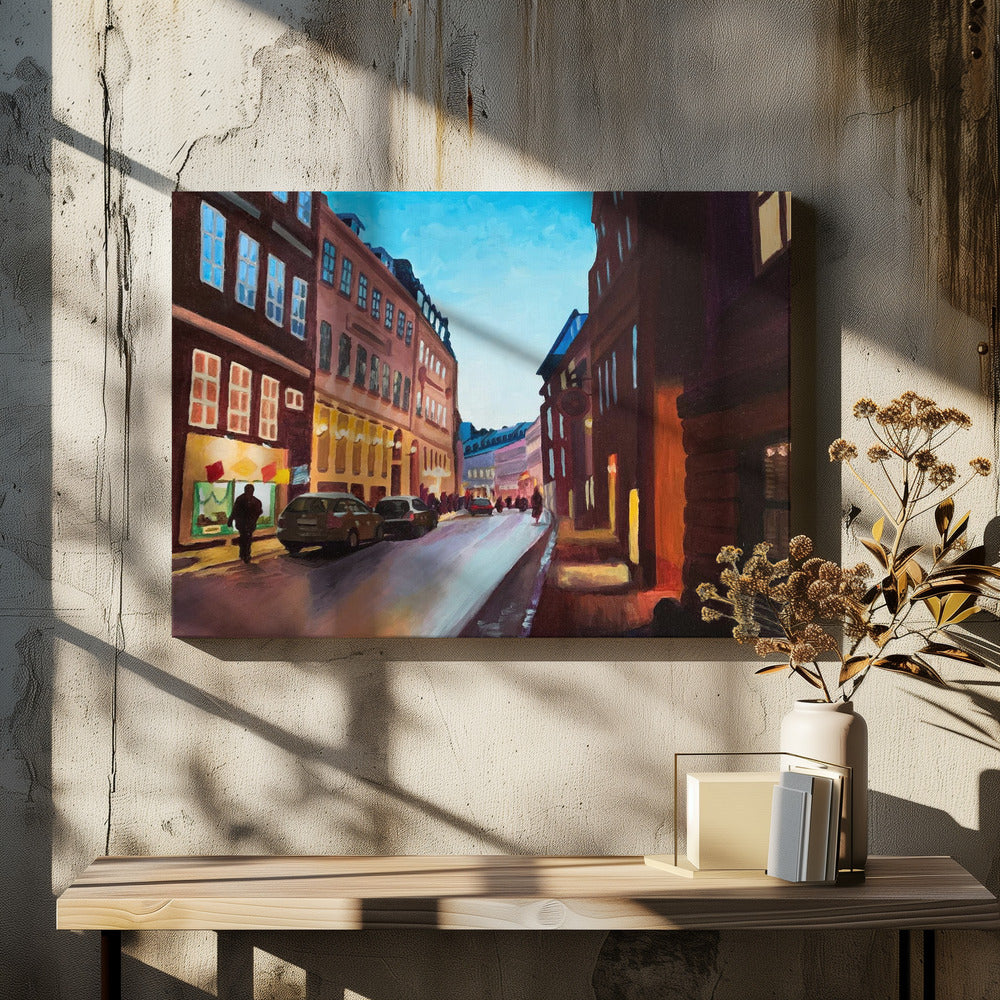 An impressionistic painting of a European city street at dusk, shown in a silver frame. The sky is a bright, clear blue, contrasting with the deep brown and maroon buildings. Warm yellow light glows from shop windows and street lamps, reflecting on the wet-looking street where cars are parked. Decor