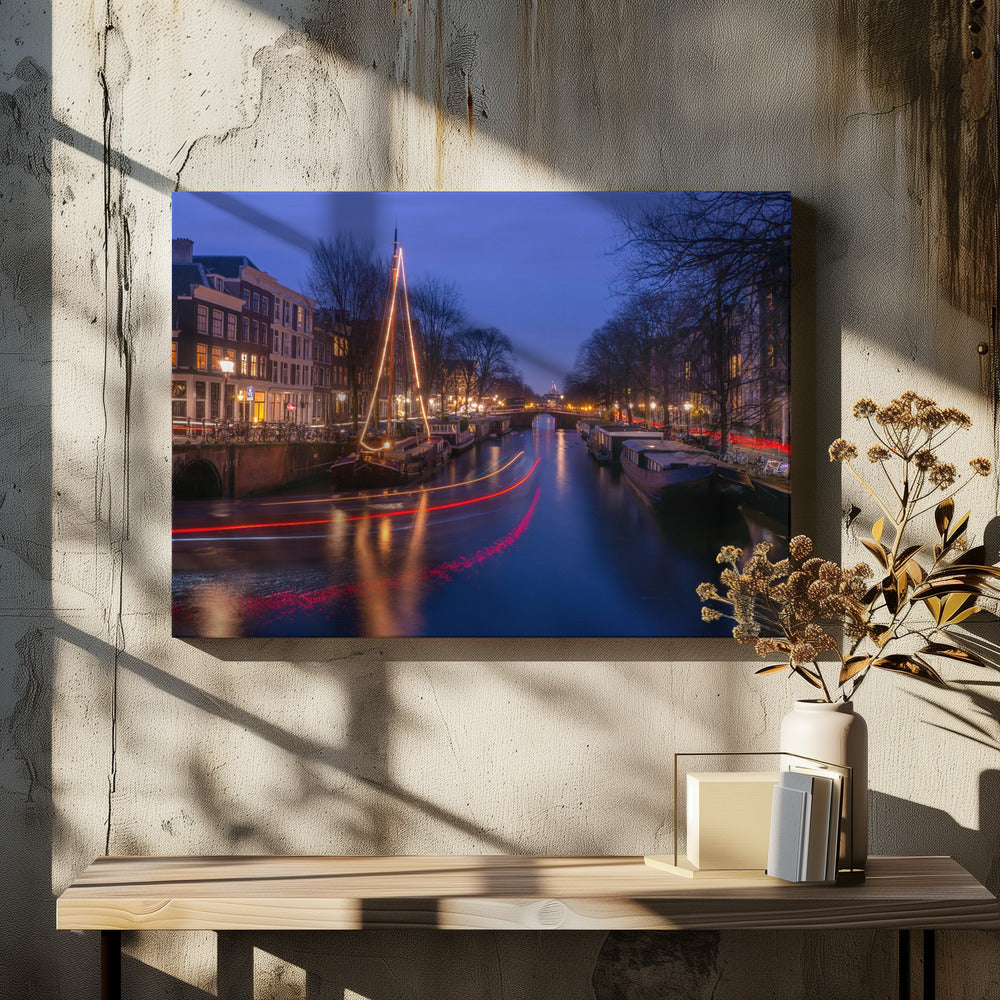 A long-exposure photograph of an Amsterdam canal at dusk. The scene is bathed in a deep blue light, with warm yellow and orange lights from street lamps and buildings reflecting on the water. A sailboat is decorated with glowing string lights, and red and white light trails from a passing boat create streaks across the canal. Houseboats are moored along the sides, and a bridge is visible in the distance. Poster