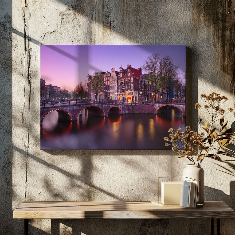A scenic view of an Amsterdam canal at twilight, with historic brick buildings and illuminated arched bridges reflected in the calm water under a vibrant pink and purple sky. Poster