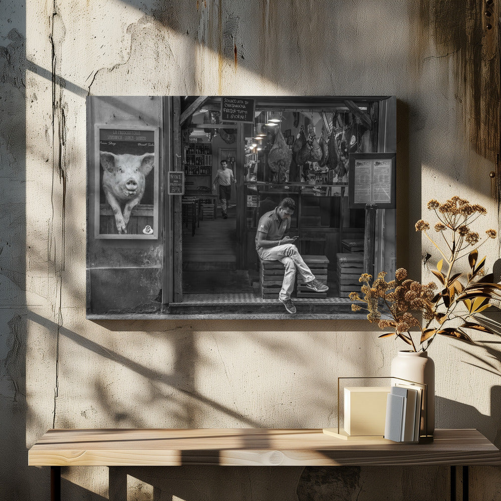 A framed, black and white street photograph of an Italian prosciutto shop. A young man sits on a wooden crate outside, looking at his phone, while cured hams hang inside the shop and a large, framed picture of a pig is on the exterior wall. Artwork