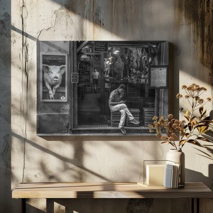 A framed, black and white street photograph of an Italian prosciutto shop. A young man sits on a wooden crate outside, looking at his phone, while cured hams hang inside the shop and a large, framed picture of a pig is on the exterior wall. Artwork