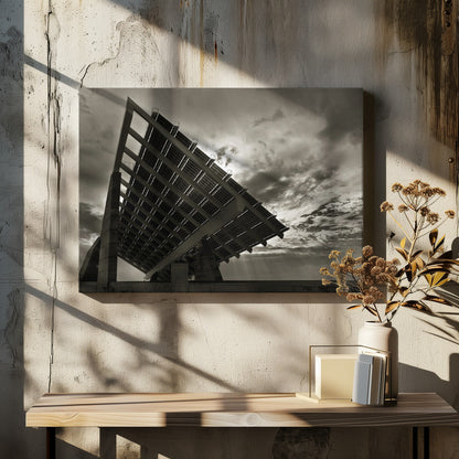 A framed, dramatic black and white photograph showing a massive, tilted solar panel structure from a low angle. The sky is filled with dark, moody clouds with some sun rays breaking through. A tiny silhouette of a person in the lower right corner emphasizes the grand scale of the architecture. Wall Art