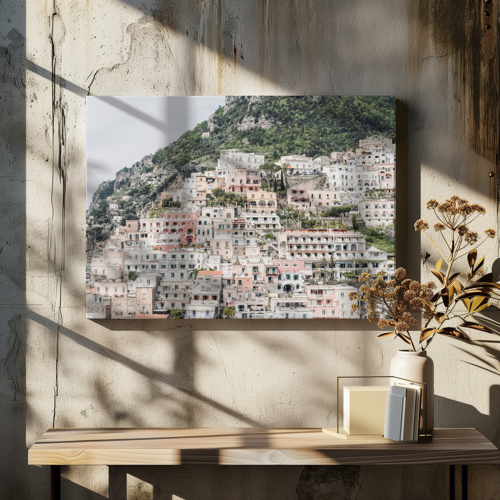 Photograph of the iconic cliffside village of Positano on the Amalfi Coast, showcasing a dense cascade of white, pink, and peach buildings down a lush green mountain under an overcast sky. The image is presented within a silver picture frame. Artwork
