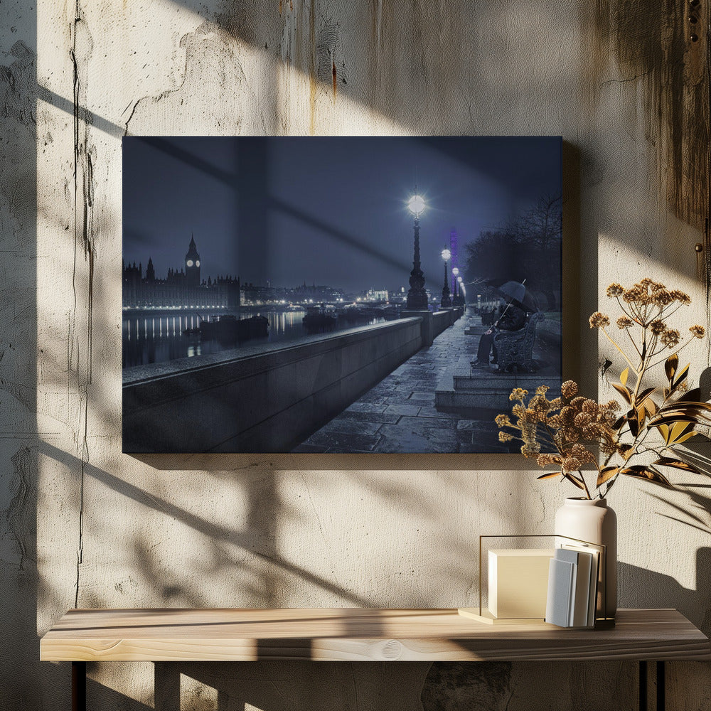 A moody, blue-toned night photograph of London. A solitary figure sits on a bench under an umbrella on a wet stone walkway along the River Thames, illuminated by vintage streetlights. Across the river, the iconic Houses of Parliament and Big Ben are lit up against the dark sky, their lights reflecting in the water. Poster