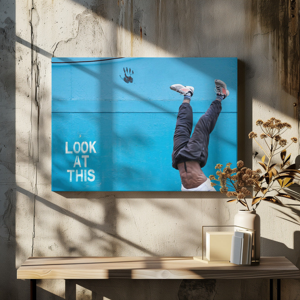 A person's lower body doing a handstand against a bright blue wall with the stenciled words 'LOOK AT THIS' in white. The person wears dark pants and sneakers, and their muscular abdomen is visible. A black handprint is on the wall above the text. The artwork is presented in a silver frame. Print