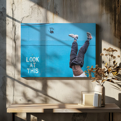 A person's lower body doing a handstand against a bright blue wall with the stenciled words 'LOOK AT THIS' in white. The person wears dark pants and sneakers, and their muscular abdomen is visible. A black handprint is on the wall above the text. The artwork is presented in a silver frame. Print