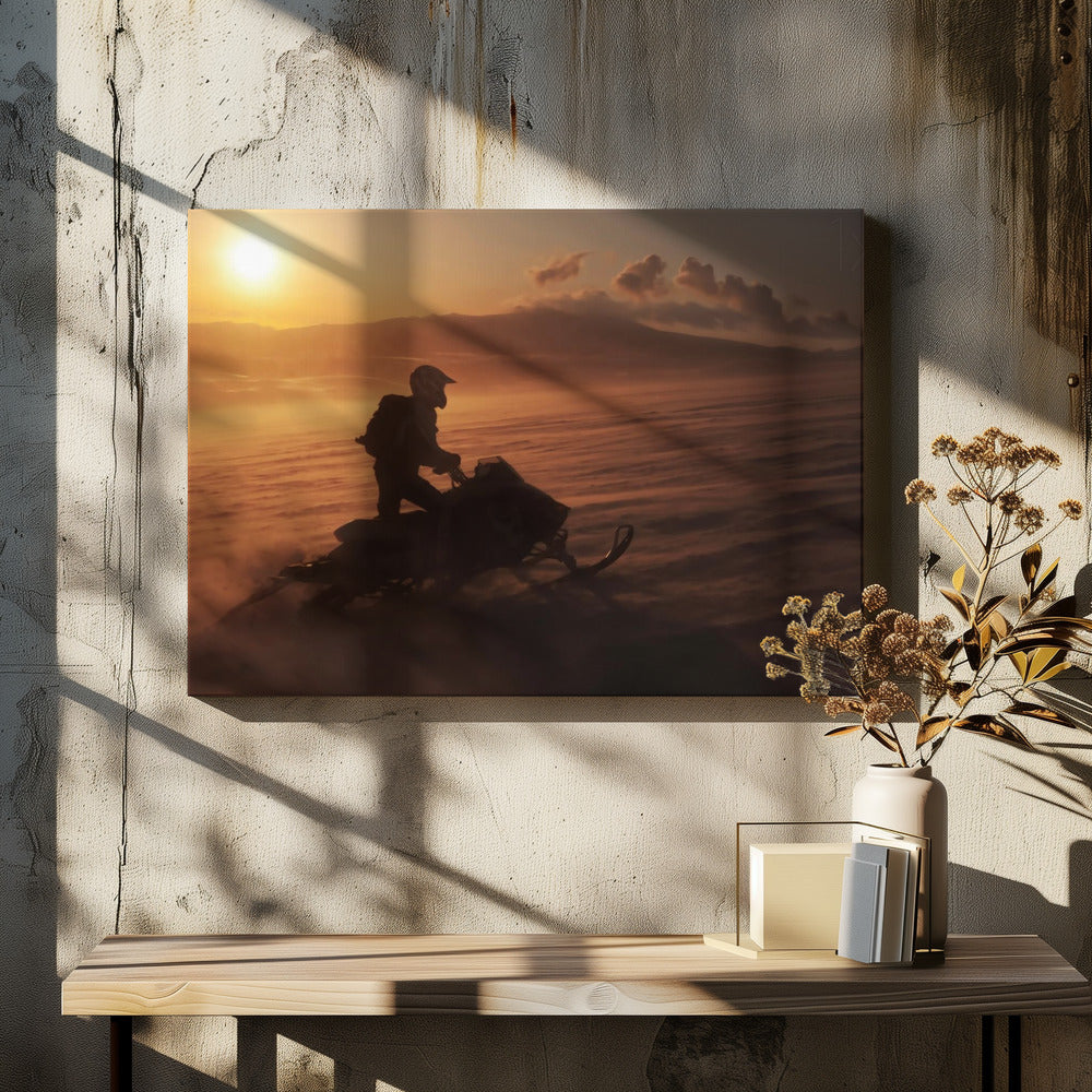A silhouette of a person wearing a helmet and backpack stands on a snowmobile in a vast, snowy landscape during a golden sunset. The wind blows snow across the ground, creating a hazy, atmospheric effect, with mountains visible in the distance under a warm sky. Wall Art