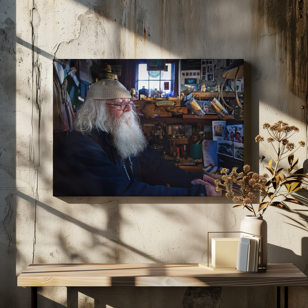 A profile portrait of an elderly man with a long, full white beard and hair, wearing glasses and a cream-colored knit beanie. He sits indoors in a warmly lit, cluttered room that looks like a cabin or a pub, with his hands clasped in front of him, gazing thoughtfully to the side. Poster