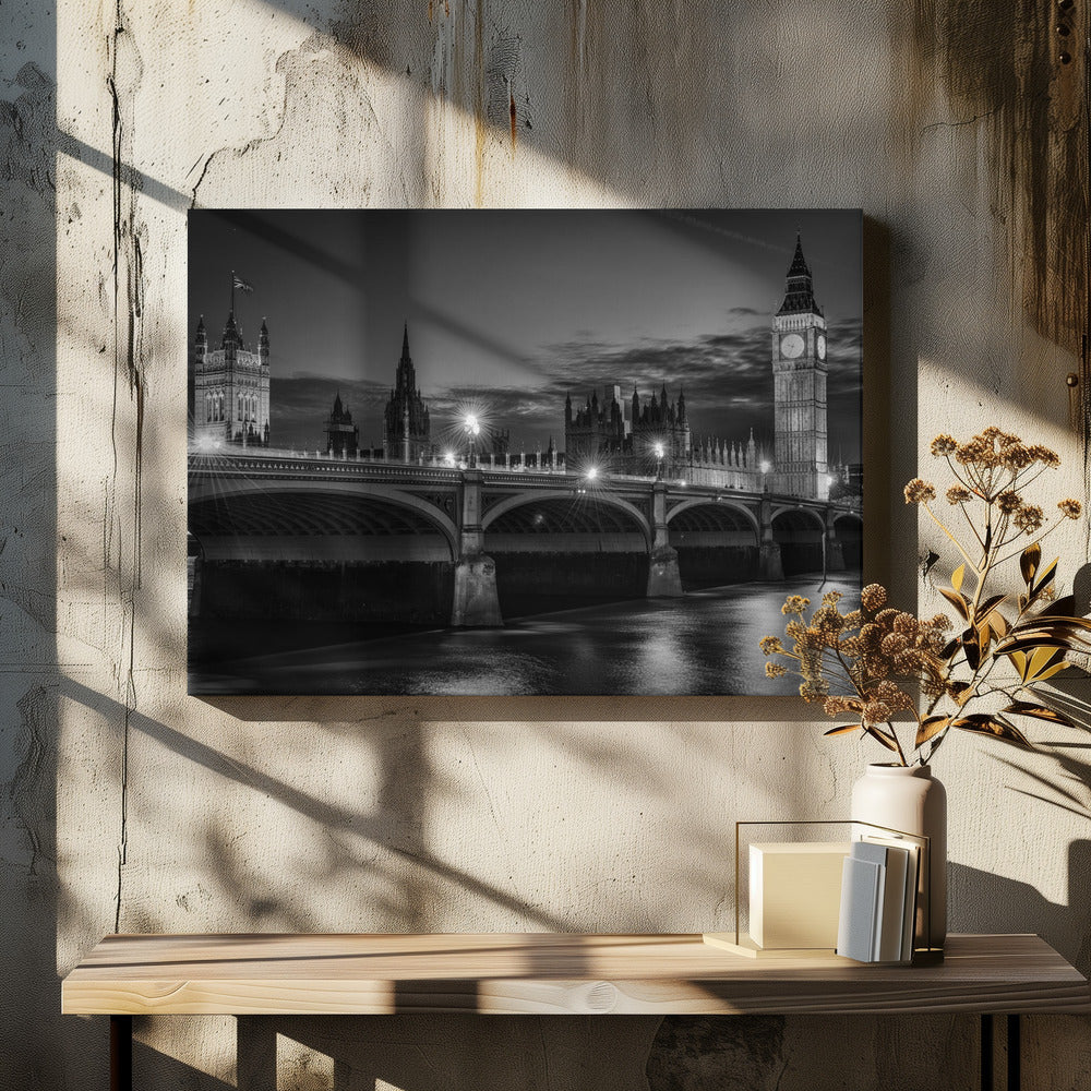 A framed, black and white nighttime photograph of Westminster Bridge in London. The iconic Houses of Parliament and the glowing Big Ben clock tower stand prominently in the background against a dark, cloudy sky. The streetlights on the bridge create starburst effects, and their reflections shimmer on the surface of the River Thames in the foreground. Poster