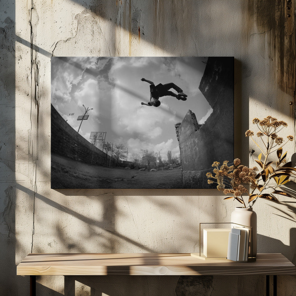 A dynamic black and white fisheye photograph of a person mid-flip, jumping over a crumbling wall in an urban setting under a cloudy sky. The image has a silver-colored frame. Print