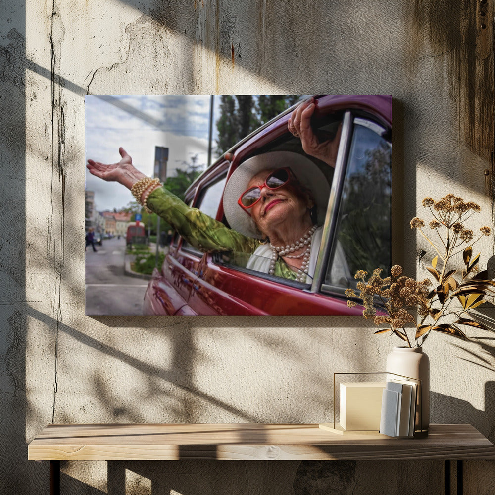 A stylish elderly woman wearing a white hat, red sunglasses, and pearl necklaces leans out of the passenger window of a classic red car. Her arm is outstretched with a joyful expression on her face, enjoying a ride through a city street. Poster