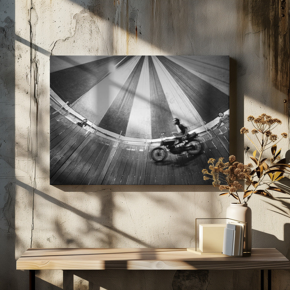 A dynamic, wide-angle black and white photograph of a motorcycle rider performing a stunt in a motordrome, commonly known as the 'wall of death'. The rider is a blur of motion as they speed along the steep, curved wooden walls. The perspective looks up towards the striped canvas top of the tent, with a few spectators visible watching from the railing at the top edge. The entire image is enclosed in a silver frame. Print