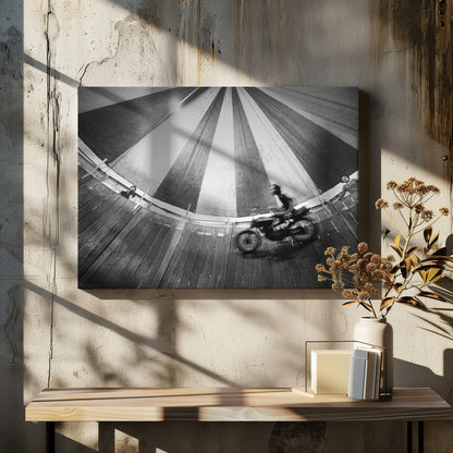 A dynamic, wide-angle black and white photograph of a motorcycle rider performing a stunt in a motordrome, commonly known as the 'wall of death'. The rider is a blur of motion as they speed along the steep, curved wooden walls. The perspective looks up towards the striped canvas top of the tent, with a few spectators visible watching from the railing at the top edge. The entire image is enclosed in a silver frame. Print