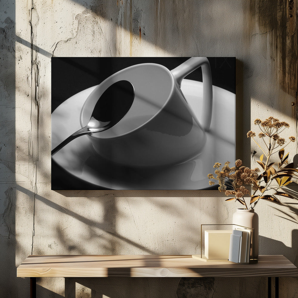 A dramatic black and white close-up photograph of a white porcelain coffee cup tilted on its saucer, with a reflective spoon resting inside. The image has high contrast against a dark background and is presented within a silver-colored frame. Decor