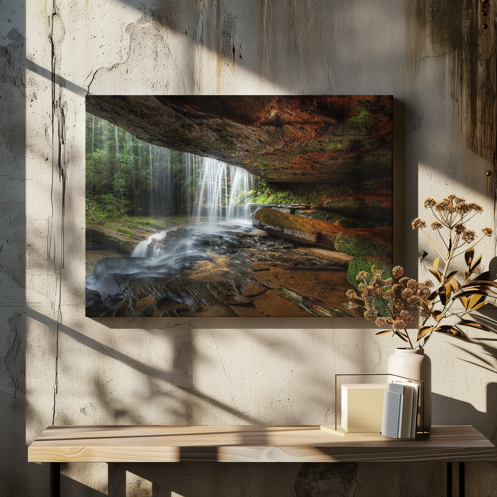 A long-exposure photograph taken from underneath a rock overhang, looking out through a cascading waterfall. The water appears silky as it flows over mossy rocks in a stream below. The scene is framed by a silver border. Print