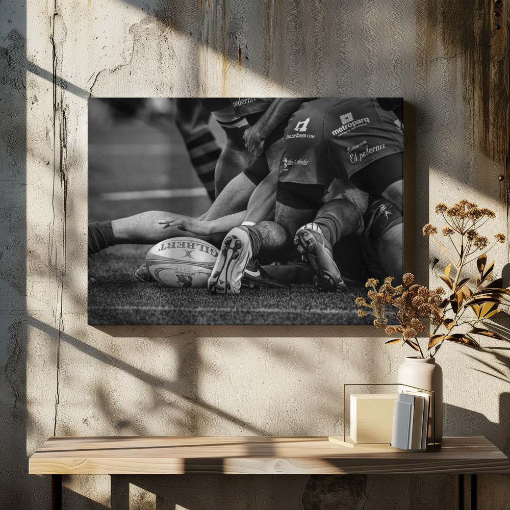 A dynamic black and white, low-angle photograph capturing the intensity of a rugby ruck. A Gilbert rugby ball sits on the turf in the foreground, surrounded by a tangle of players' muscular legs, arms, and cleats, conveying the raw power and action of the sport. Wall Art