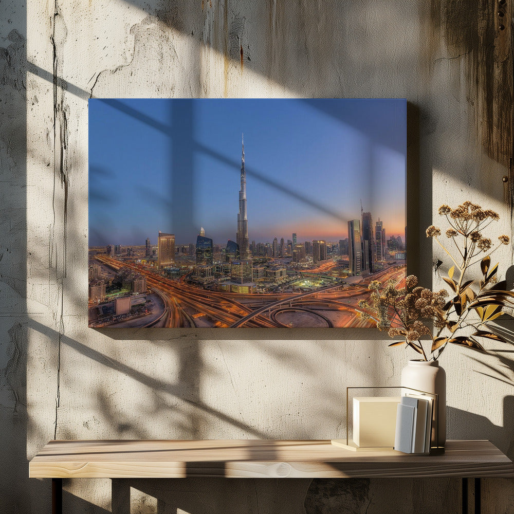 An aerial photograph of the Dubai skyline at dusk, centered on the illuminated Burj Khalifa. The sky transitions from deep blue to warm orange at the horizon, while a complex network of highways below glows with the light trails of traffic. Artwork
