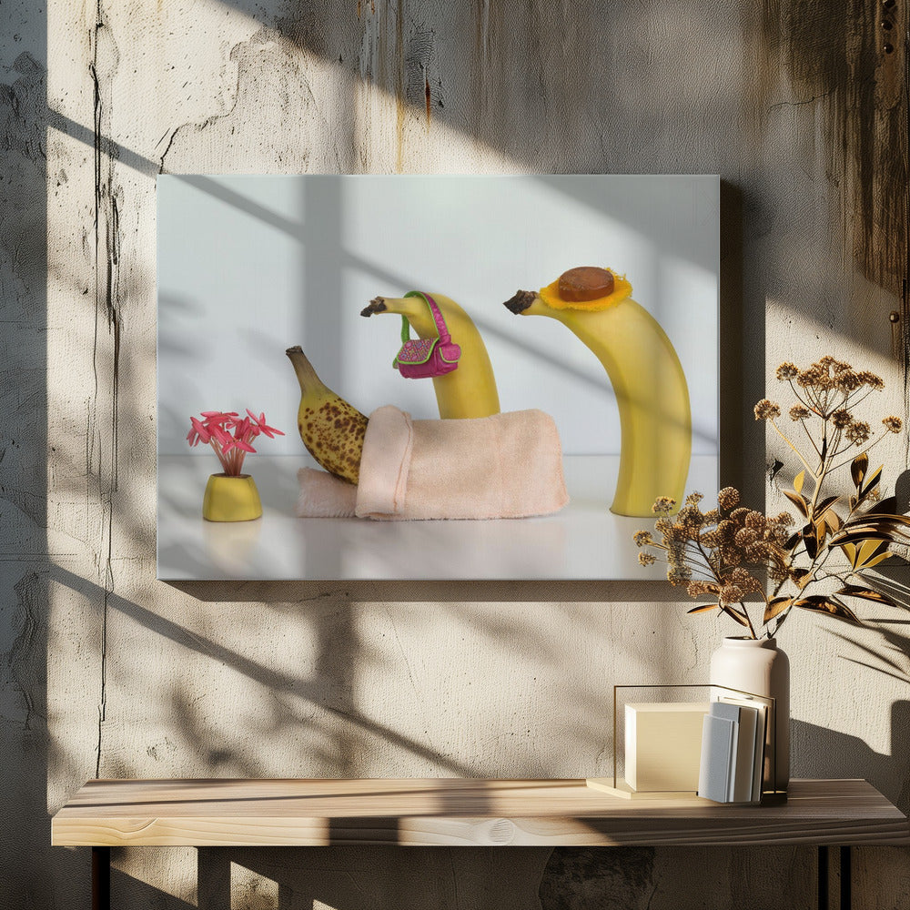 A whimsical still life photo featuring three bananas personified as a family or friends. One overripe, spotted banana lies down, covered by a pink towel, while another banana with a tiny pink purse and a third with a small hat seem to be visiting. A small vase with pink flowers sits nearby on the white surface. Poster
