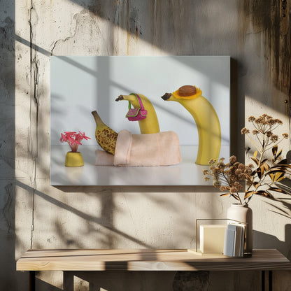 A whimsical still life photo featuring three bananas personified as a family or friends. One overripe, spotted banana lies down, covered by a pink towel, while another banana with a tiny pink purse and a third with a small hat seem to be visiting. A small vase with pink flowers sits nearby on the white surface. Poster