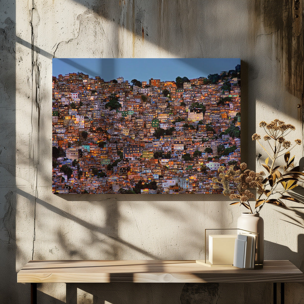 A photograph of a densely packed favela built on a hillside at dusk. The numerous small, colorful houses are illuminated from within, casting a warm orange glow that contrasts with the deep blue of the twilight sky. The image is presented within a silver picture frame. Artwork