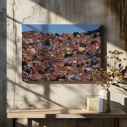 A photograph of a densely packed favela built on a hillside at dusk. The numerous small, colorful houses are illuminated from within, casting a warm orange glow that contrasts with the deep blue of the twilight sky. The image is presented within a silver picture frame. Artwork