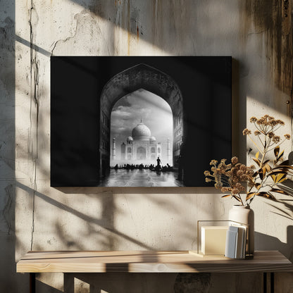 A dramatic black and white photograph of the Taj Mahal seen through a large, dark archway which frames the view. The iconic mausoleum is in the distance under a cloudy sky, and the silhouettes of many people are scattered on the wet, reflective ground in the foreground. Decor