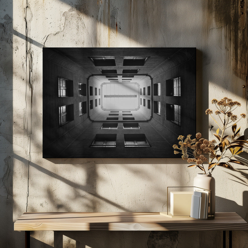 A symmetrical, black and white photograph looking directly up from the base of a building's interior courtyard. The dark walls rise up, lined with rows of windows, creating a powerful sense of perspective that draws the eye to the bright rectangle of the sky or a skylight at the top. The image is enclosed in a simple silver frame. Poster