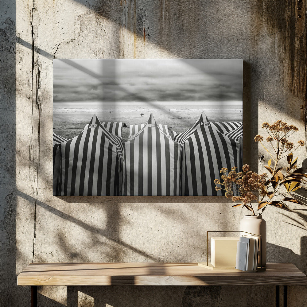 A framed, black and white photograph of striped beach tents on a cloudy day, with the ocean and distant surfers in the background. Print