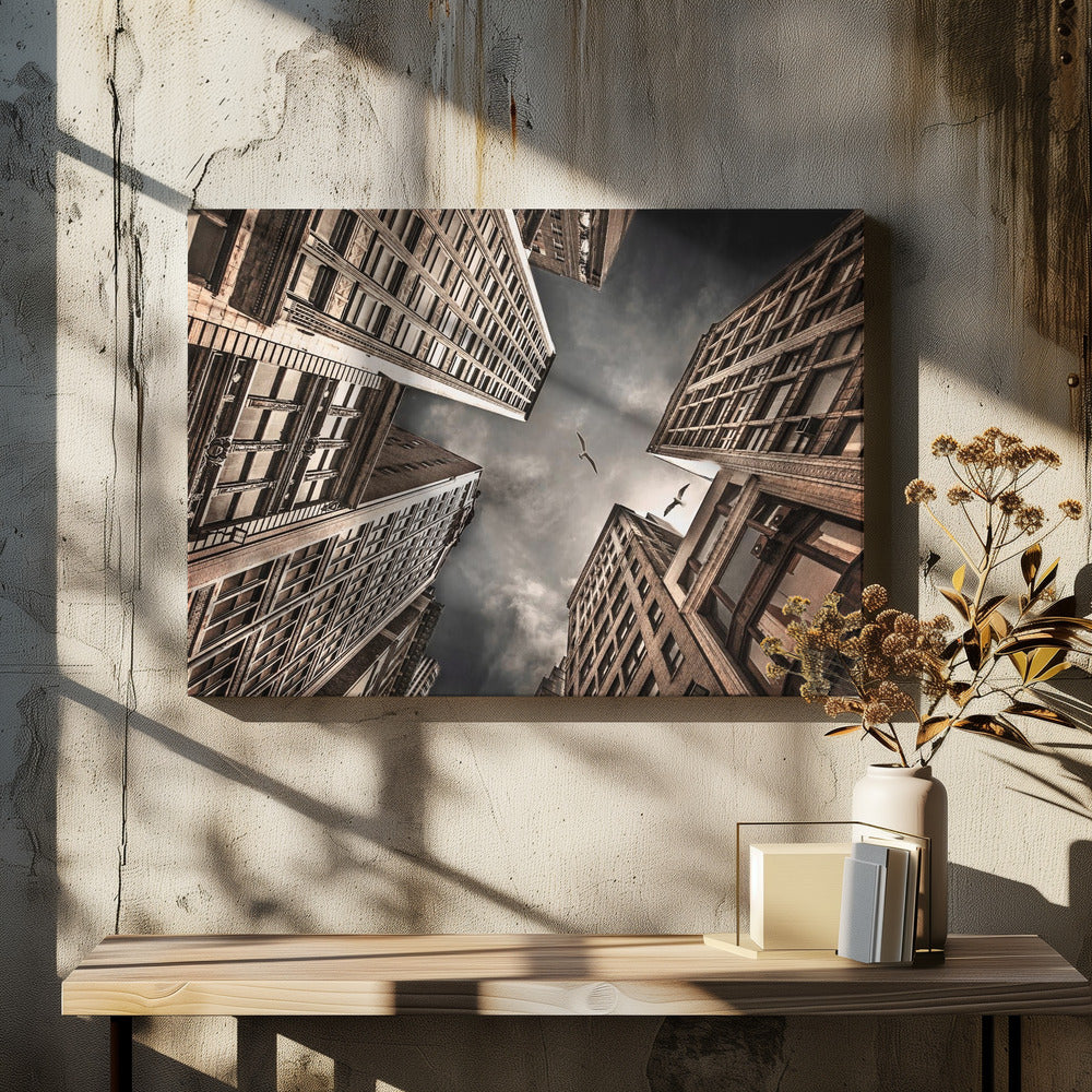 A dramatic, low-angle photograph looking up at the sky between several tall, sepia-toned skyscrapers that converge towards the center. Two birds are seen flying in the moody, dark grey sky. The image has a vintage feel and is presented in a silver frame. Wall Art