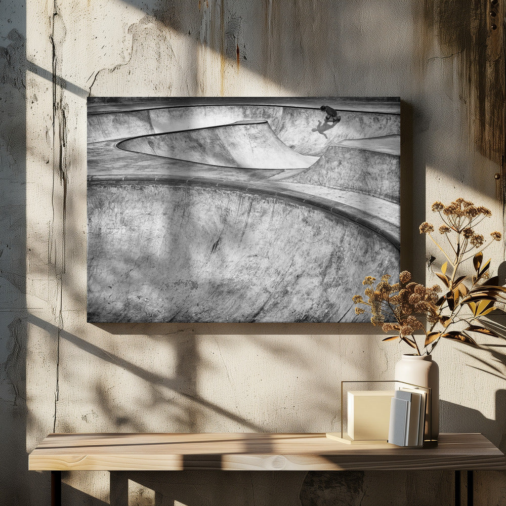 A high-angle, black and white photograph of a sprawling concrete skatepark with smooth, curving ramps. A single skateboarder is visible in the upper right, casting a distinct shadow on the ramp as they ride. The image is enclosed in a silver frame. Decor