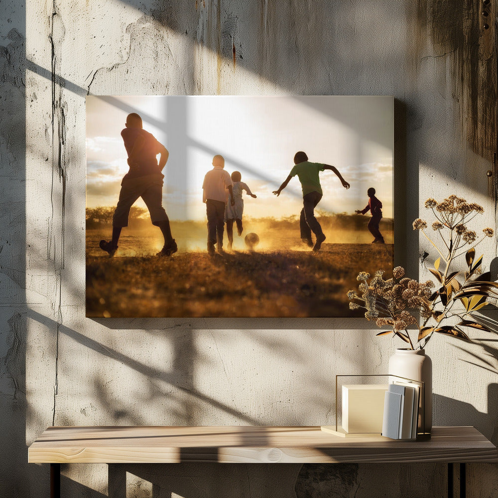 A low-angle, warmly lit photograph of five children silhouetted against a bright sunset as they play soccer on a dusty field, kicking up clouds of dust in the golden light. Decor