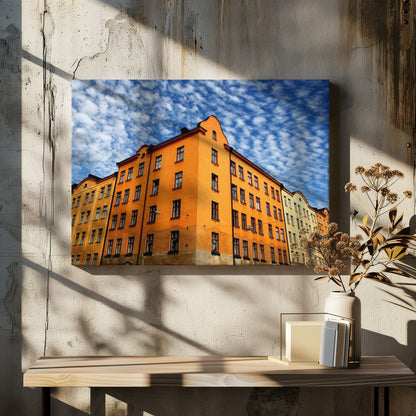 A low-angle shot of a vibrant orange apartment building against a dramatic blue sky filled with small, puffy white clouds. The building is on a corner, with another yellow-hued building visible next to it. The entire image is enclosed in a silver, brushed-metal style frame. Artwork