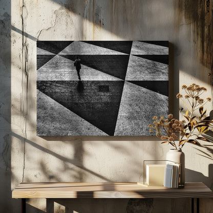 A dramatic, high-contrast black and white overhead photograph of a person walking across a paved ground with a striking geometric pattern of light and dark triangles created by strong shadows. The lone figure casts a long shadow, enhancing the minimalist and abstract composition, which is enclosed in a grey frame. Print