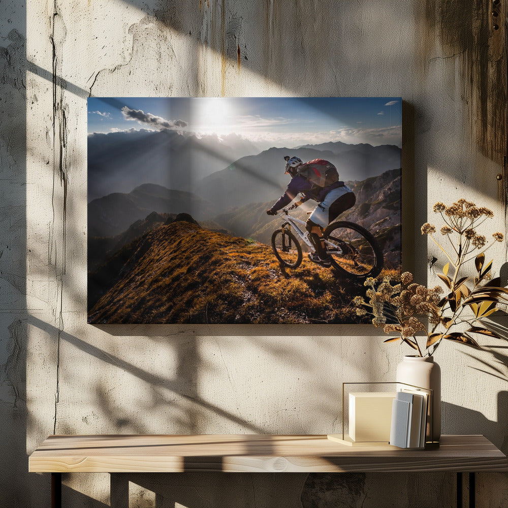 A mountain biker navigates a narrow, golden-lit ridge against a backdrop of a vast mountain range. The sun is breaking through clouds in the distance, casting dramatic rays of light across the epic landscape. Print