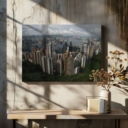 A high-angle photograph of the Hong Kong cityscape, viewed from a lush green peak. Numerous skyscrapers fill the frame, overlooking a bustling harbor with mountains in the distant background under a cloudy sky. The entire image is presented within a silver, brushed-metal frame. Artwork