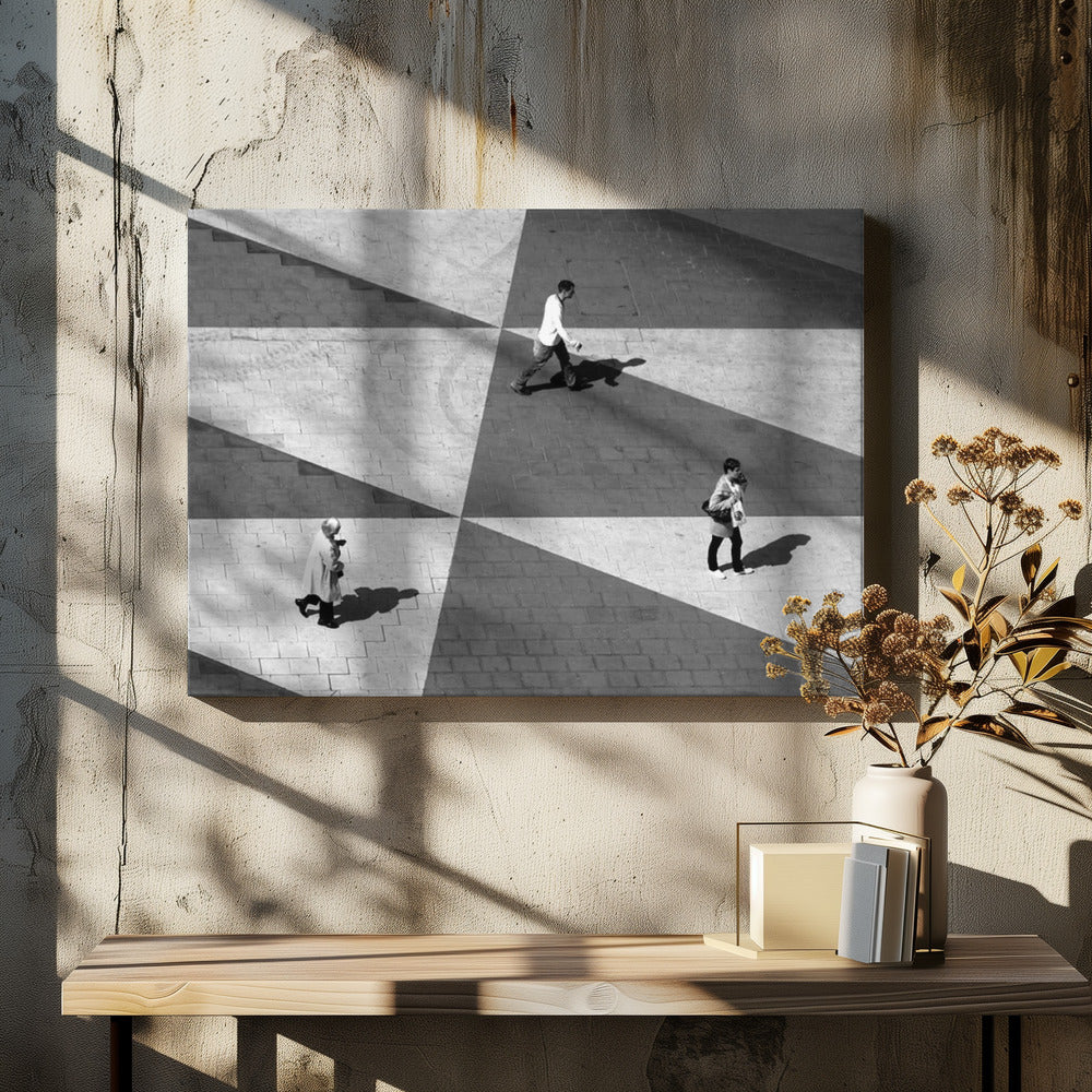 A high-angle, black and white photograph capturing three people walking across a paved plaza, their paths intersecting with a dramatic geometric pattern of light and shadow. The strong sunlight creates large, sharp-edged triangles of shade, isolating each figure in the composition, which is enclosed in a silver frame. Decor