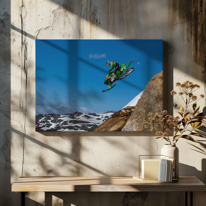 A dynamic action shot of a person in full gear launching a green snowmobile into the air off a rocky, snow-covered cliff, set against a backdrop of a vivid blue sky and distant snowy mountains. The image is enclosed in a silver frame. Wall Art