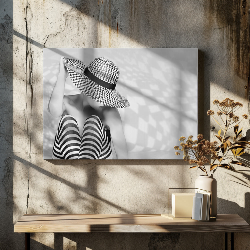 A black and white artistic photograph of a woman sitting down, her face obscured by a large, wide-brimmed sun hat with a black and white chevron pattern. She is wearing black and white striped leggings and holding the brim of her hat with one hand. The image is set against a plain background with patterned shadows and is presented within a silver frame. Artwork