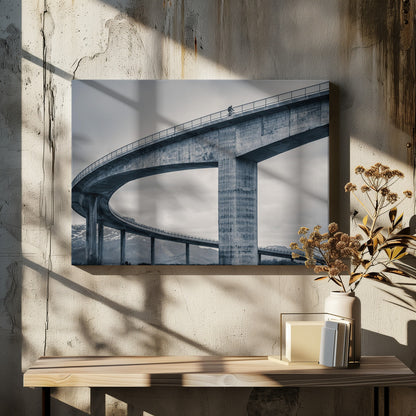 A black and white photograph of a lone cyclist riding across the top level of a massive, curving concrete bridge, which sweeps across the frame. In the background, a lower level of the bridge and distant mountains are visible under a cloudy sky. Decor