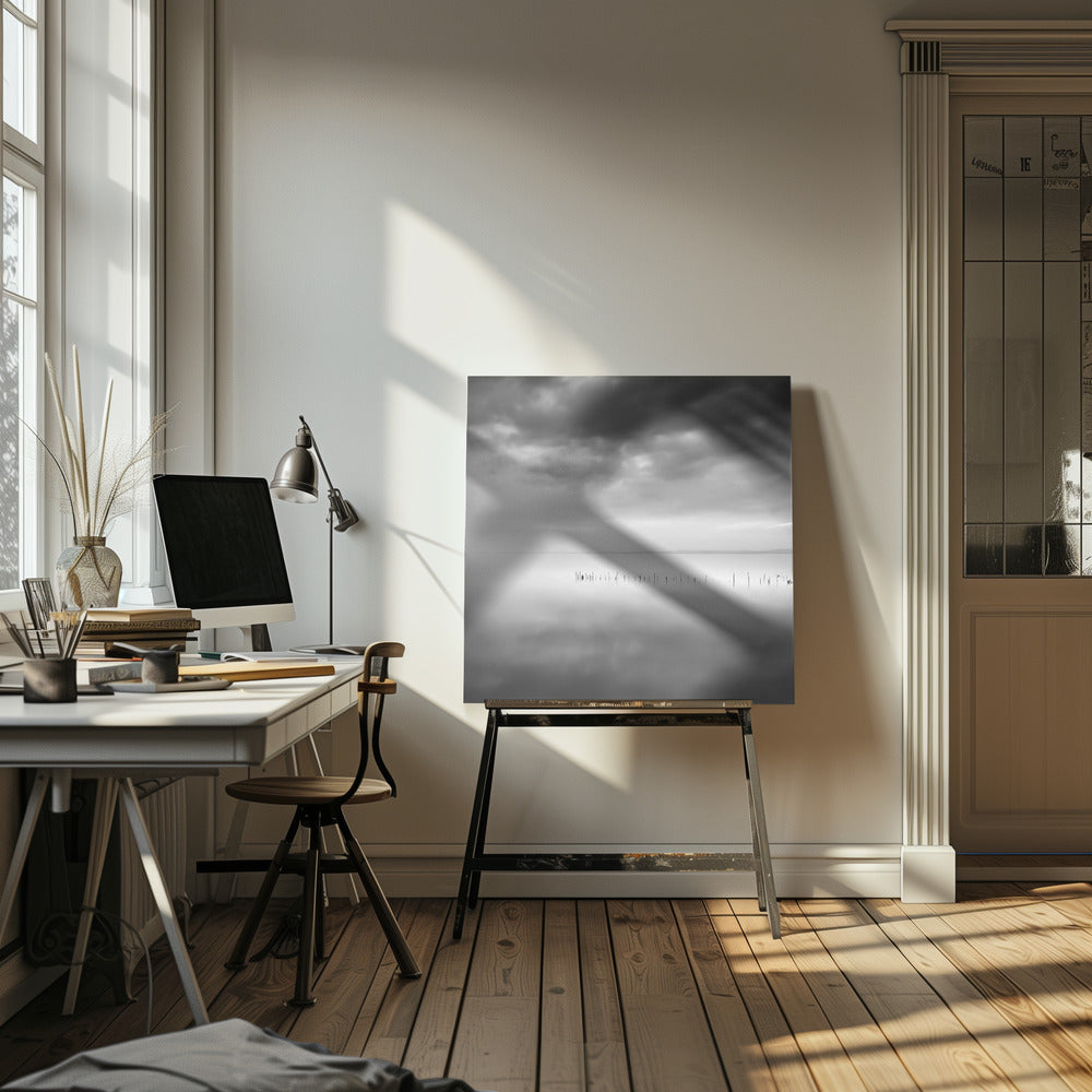 A minimalist black and white photograph in a light wood frame, depicting a calm, serene lake under a heavy, cloudy sky. The still water reflects the sky, with a row of weathered wooden posts breaking the surface along the horizon. Print
